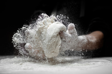 White flour flies in air on black background, pastry chef claps hands and prepares yeast dough for pizza pasta