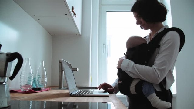 Mother Using Laptop on Kitchen Counter with Baby Sleeping in Sling