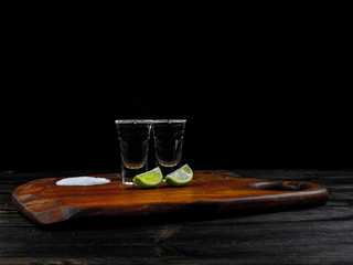 Two tequila silver shots with fresh lime and sea salt on wooden board and dark background