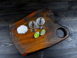 Two tequila silver shots with fresh lime and sea salt on wooden board and dark background. view from above