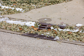 Storm sewer gutter drain grate in city street curb covered in melting snow and leaves after autumn...