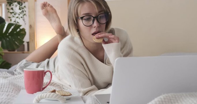 Relaxed woman in pajamas using laptop and eating biscuit in bed