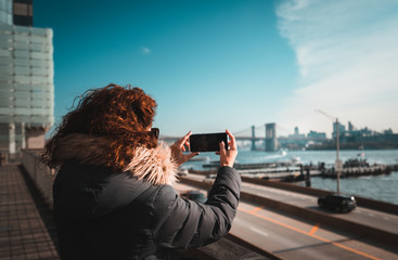 Beautiful curly brunette woman taking selfie self-portrait with the Brookling bridge on the background while sightseeing new york during winter season