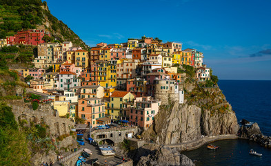 Fototapeta na wymiar Manarola typical Italian village in National park Cinque Terre, colorful multi colored buildings houses on rock cliff, fishing boats on water, blue sky background, Liguria, Italy