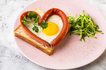 Scrambled eggs in the form of heart on a white plate with sausages, sourdough toast isolated on white marble background. Homemade food. Tasty breakfast. Selective focus. Hotizontal photo.