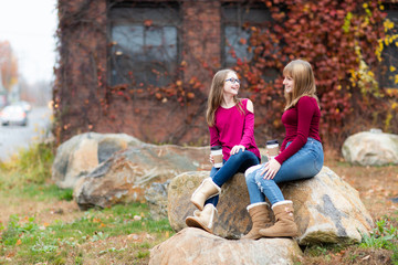 Two young teenager girls sitting on the rock, drinking coffee. Sisters, friends happy smiling, laughing, talking. Fall autumn time.Playing clapping hands 