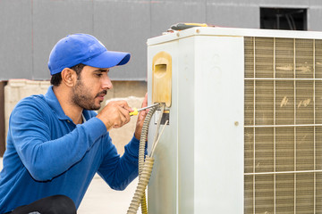a professional electrician man is fixing a heavy duty unit of central air conditioning system by his tools on the roof top and wearing blue color of uniform