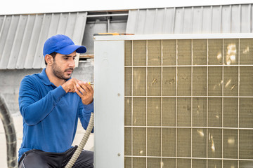 a professional electrician man is fixing a heavy duty unit of central air conditioning system by his tools on the roof top and wearing blue color of uniform