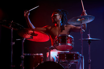 Fototapeta premium Portrait of active african man playing on drums set in neon lights, wearing eyeglasses, performing music. expressive emotions and movement