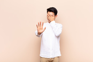 young chinese man covering face with hand and putting other hand up front to stop camera, refusing...