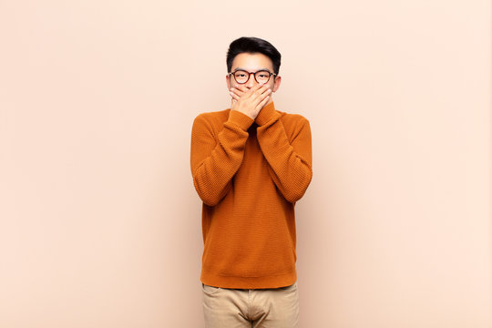 young chinese man covering mouth with hands with a shocked, surprised expression, keeping a secret or saying oops against flat color wall