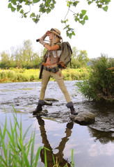 A young girl dresses up as an explorer. She wears a safari hat and wears khaki clothing. She is seen in a  river environment.