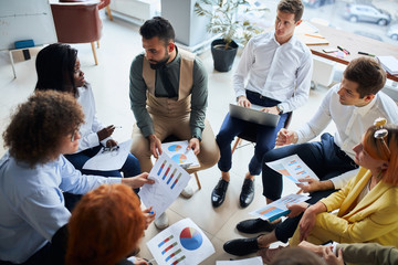 Modern and enthusiastic business people sit in circle discussing business ideas, steps for building business, using diagrams and papers.