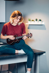 Fototapeta na wymiar portrait of nice young caucasian woman wearing casual clothes sit on table playing ukulele, indoors. look at instrument