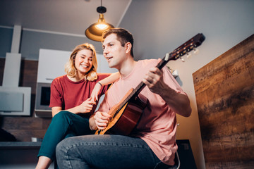 caucasian young male in casual wear sit with her blond short-haired girlfriend playing guitar for her and singing