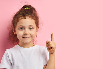 Smiling attractive girl kid show forefinger, wearing white t-shirt, isolated pink background