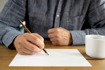 The man is holding a pen over a blank white sheet. Front view on the background of a wooden table.