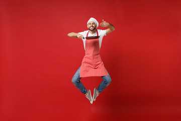 Smiling young bearded male chef cook or baker man in striped apron white t-shirt toque chefs hat posing isolated on red background. Cooking food concept. Mock up copy space. Jumping, hold frying pan.