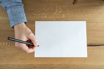 The man is holding a pen over a blank white sheet. Top view on the background of a wooden table.