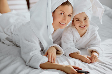 happy caucasian mother and her little child girl dressed in bathrobe and towel looking at camera and holding tv remote