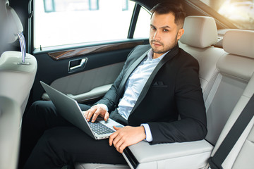 handsome young businessman sit with laptop while working in car, man looking at camera