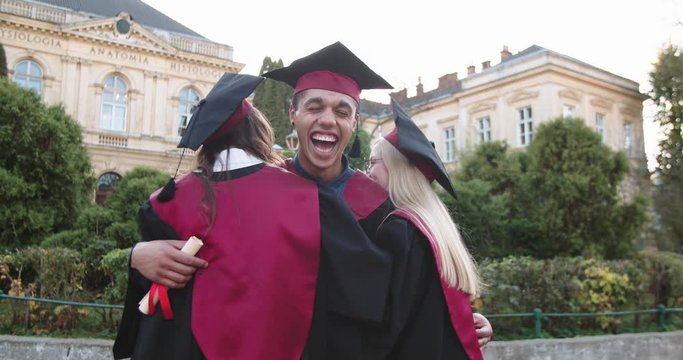 Portrait shot of the young Caucasian cheerful two girls hugging their friend - moulatto guy, happy master graduated students smiling joyfully to the camera after graduation ceremony. Outdoor.