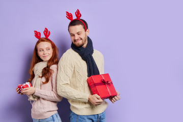 Attractive delightful woman in warm comfortable sweater ready to exchange Christmas gifts with handsome caucasian man, standing with backs to each others, turning heads back, Merry Christmas