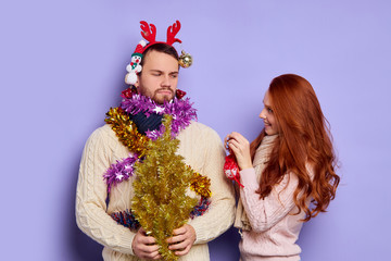 Cute funny man looking at pretty redhead female friend with displeased expression, young lady putting Christmas tinsel and different baubles on angry man, kidding and making jokes, studio shot