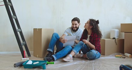 Caucasian young and cheerful man and woman talking and deciding how to decorate a room and colour of walls while having repair at home and sitting on the floor at the boxes. - Powered by Adobe