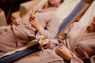 Young caucasian couple take care of body together lying in spa salon and drinking champagne, wearing bathrobe