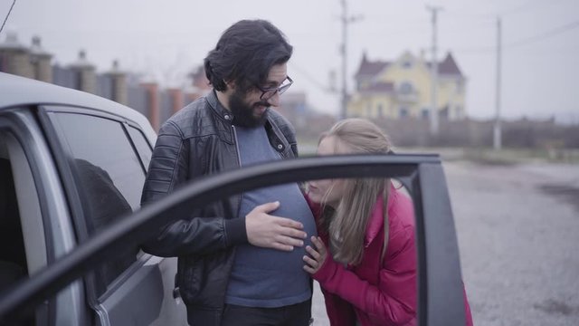Positive bearded Caucasian man in eyeglasses imitating baby hitting as her girlfriend or wife holding ear next to his pregnant belly. Funny family exchanging their social roles. Joking, fun.