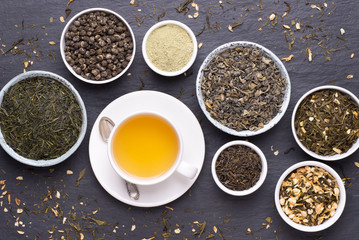 Cup of green tea and bowls of various dried tea leaves on dark, stone background, top view