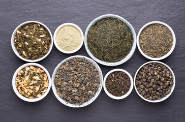 Assortment of dried tea leaves in bowls on dark, stone background, top view