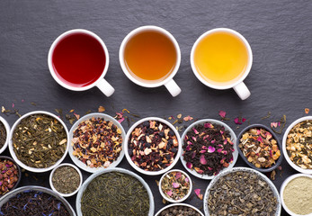  Assortment of tea cups, dried leaves, fruit and herbs in bowls on dark, stone background, top view