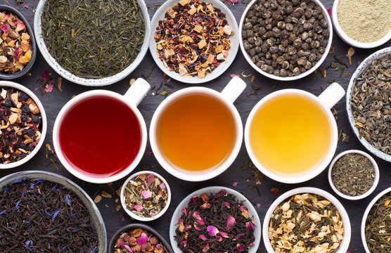  Assortment Of Tea Cups, Dried Leaves, Fruit And Herbs In Bowls On Dark, Stone Background, Top View