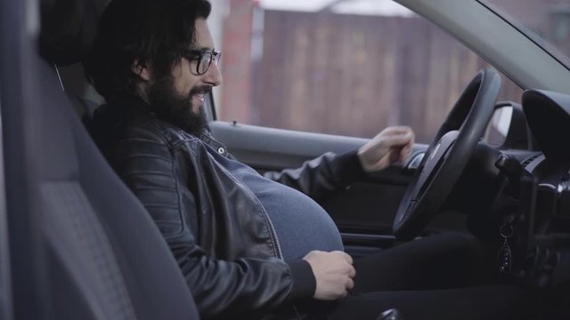 Side view close-up of funny pregnant man sitting at driver's seat and caressing his belly. Portrait of smiling adult guy with curly black beard wearing eyeglasses. Joking, fun.