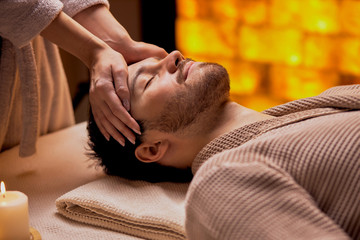 Obraz na płótnie Canvas Spa, treatment concept. Young male wearing bathrobe lying on table with closed eyes and get massage by woman's hands.