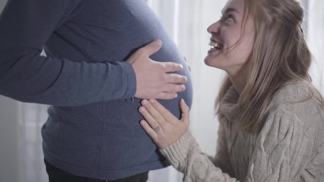 Portrait of smiling Caucasian woman kissing male's pregnant belly and listening to baby's heartbeat. Funny couple exchanging their social roles. Joking, fun, kidding.