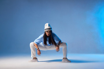 Attractive young dancer with long straight brunette hair, dressed in light coloured clothes, sitting on squat, putting hands on knees, looking at camera ready to start performance, blue smoke