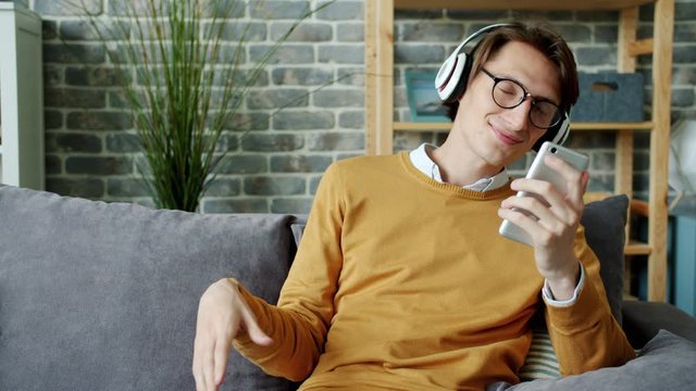 Slow motion of cheerful young man melomaniac enjoying music throught wireless headphones and using smartphone. People, lifestyle and gadgets concept.