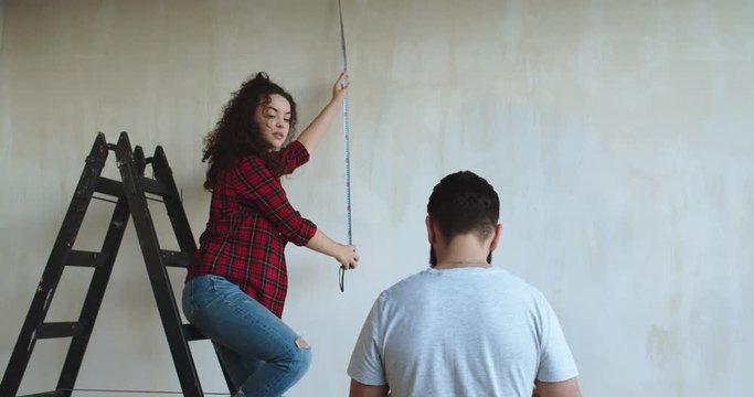Caucasian pretty woman standing on the ladder in the room measuring wall with a measure tape and her husband coming to her with a tablet devise as showing some ideas of renovation on it.