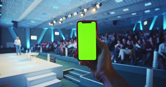 Close up of young man hand scrolling blank green screen smart phone mobile with crowded evening conference hall and speaker on stage on background. Chroma key display. Tutorials seminars online.