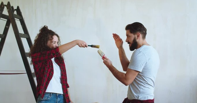 Caucasian young happy and cheerful couple playing, dancing, fighting with brushes while having fun duren renovation in the apartment.