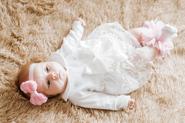 A three-month-old girl lies on a brown blanket wearing a white dress and a pink headband. Photo from above.