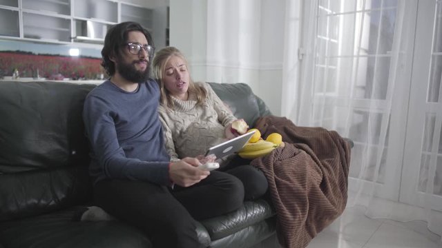 Young Caucasian couple sitting on the couch at home and watching TV. Bearded man in eyeglasses switching channels and his beautiful pregnant wife eating apple and looking at tablet screen. Gestation.