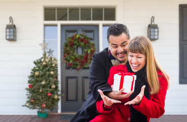 Young Mixed Race Couple Exchanging Gift On Front Porch of House with Christmas Decorations