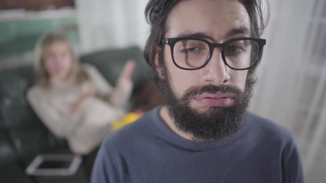 Close-up portrait of young Caucasian man with black curly beard wearing eyeglasses. Blond pregnant woman yelling at him from the background. Pregnancy, gestation, mood swings, patience.