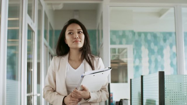 Attractive asian businesswoman holding documents and walking through the office towards the camera