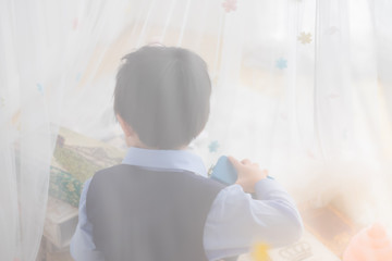 Obraz na płótnie Canvas An Asia little boy who wear navy suit and bow tie sitting back behind white sheer curtains which decorated with star