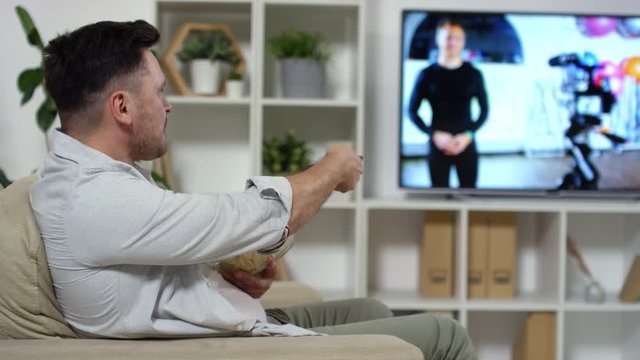 Middle aged Caucasian man sitting in comfortable armchair, eating potato chips from glass bowl and using remote control while watching TV in the living room; focus in the background